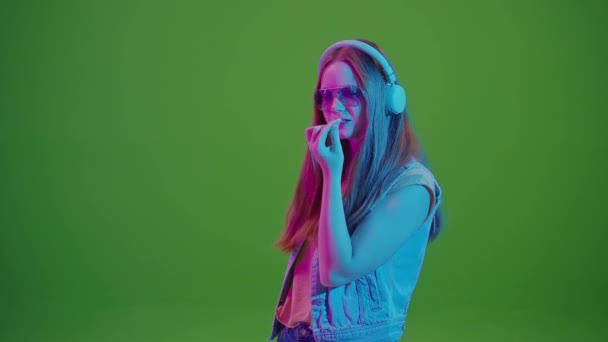 グリーン・スクリーン。 ネオンライトで踊るティーンエイジャーガール, 彼女のヘッドフォンから音楽に没頭, 大きなピンクバブルガムを噛みます. 現代の若者文化と音楽への情熱のレトロバイブの融合. - 映像、動画