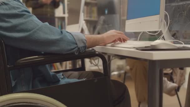 Střední záběr zblízka na ruce a střed nerozpoznatelného muže se zdravotním postižením, který sedí na invalidním vozíku před stolním počítačem v univerzitní knihovně a píše na klávesnici - Záběry, video