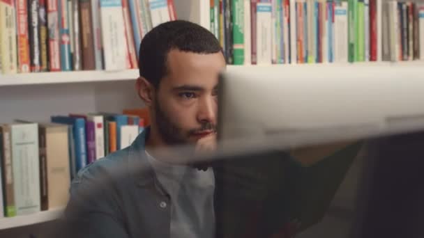 Střední detailní záběr zaměřeného mladého středovýchodního studenta, který sedí u stolního počítače v knihovně, dívá se na obrazovku, přemýšlí a píše na klávesnici, zatímco pracuje na diplomové práci - Záběry, video