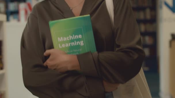 Detailní záběr pomalého záběru rukou a střední části nepoznatelné mladé studentky procházející knihovnickým sálem, svírající učebnici strojového učení a nervových sítí - Záběry, video