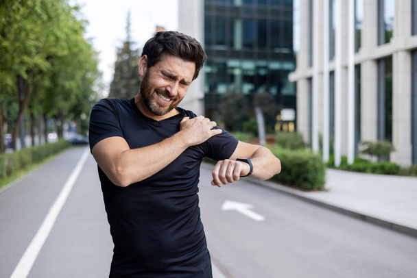 Man in zwart shirt ervaren schouder pijn tijdens het joggen in stedelijke omgeving, grimmig, controleren smartwatch. - Foto, afbeelding