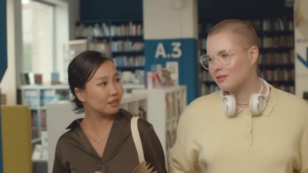 Střední stopa záběru dvou multietnických studentek v neformálních šatech procházejících společně veřejnou knihovnou, asijské ženy držící knihu, diskutující univerzitní život a usmívající se - Záběry, video