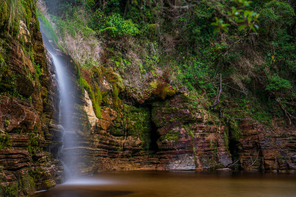 Spokojne zdjęcie długiej ekspozycji rejestruje wodospad wśród bujnych liści i kamieni. - Zdjęcie, obraz