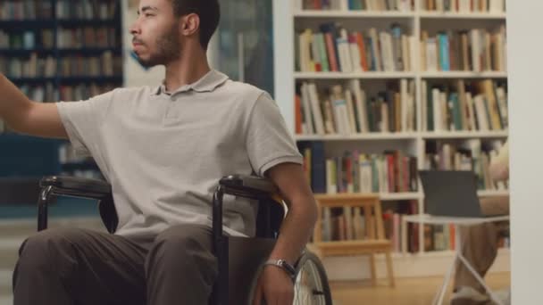 Medium tilting shot of young Arab guy with disability, in wheelchair rolling around public library, browsing through books on display on shelves, looking at titles and choosing literature for study - Footage, Video