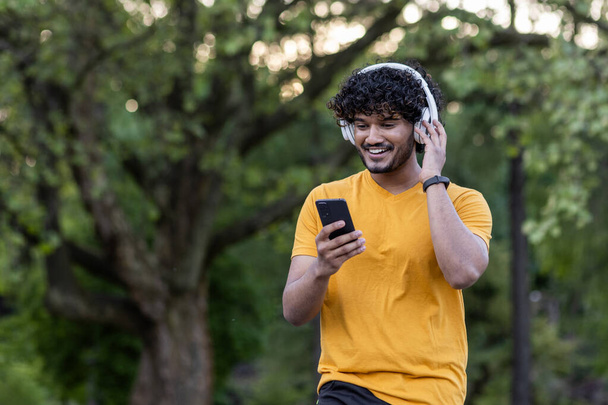 Indiański uśmiechnięty młodzieniec uprawiający sport i jogging w parku, noszący słuchawki i korzystający z telefonu komórkowego. - Zdjęcie, obraz