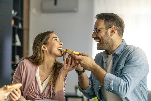 Evde pizza paylaşan genç ve güzel bir çiftin fotoğrafını çek. Genç adam ve mutlu kadın pizzayı paylaşıyor ve birlikte yiyorlar.. - Fotoğraf, Görsel