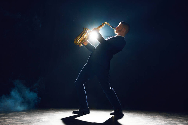 Tyylikäs mies saksofoni kallellaan ylöspäin suorittaa uuden melodian lavalla vastaan tumma tausta teatteri savua. Taiteen, instrumentaalimusiikin, tanssin, kulttuurin, festivaalien ja konserttien käsite. - Valokuva, kuva