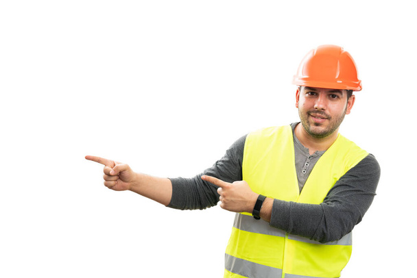 Veselý muž konstruktér s úsměvem ukazuje na prázdné kopírovací prostor pro reklamu pomocí ukazováčků na sobě pracovní oděv vesta a oranžová bezpečnostní přilba izolované na bílém pozadí - Fotografie, Obrázek
