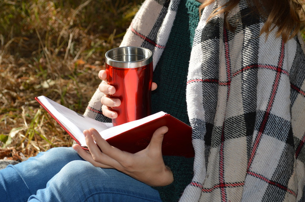 Une jeune et belle fille enveloppée dans une couverture chaude à carreaux buvant du thé chaud et lisant un livre dans le parc
 - Photo, image