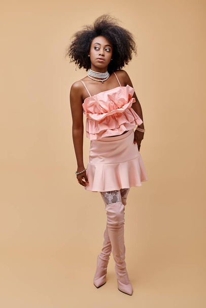 dreamy african american girl in 20s posing in peach ruffle top and over-knee boots on beige backdrop - Photo, Image