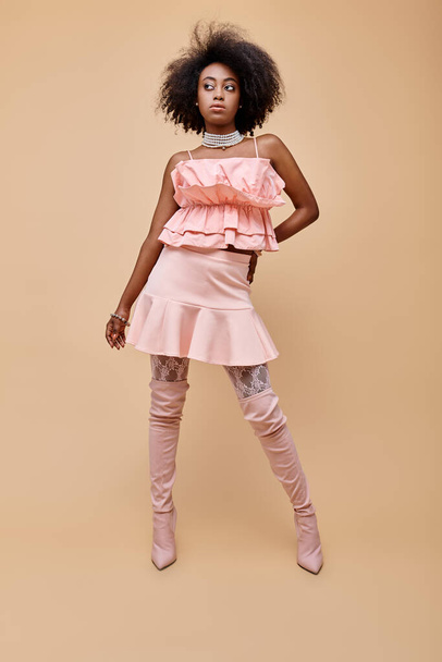 fancy african american woman in 20s, posing in peach ruffle top and over-knee boots on beige - Photo, Image
