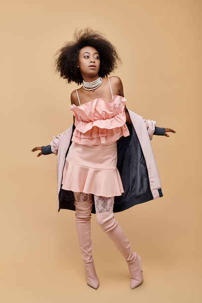 young african american woman in 20s, posing in peach outfit with jacket and over-knee boots on beige - Foto, Bild