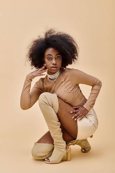 curly african american model in trendy peach fuzz outfit and thigh-high boots sitting on a beige - Photo, Image