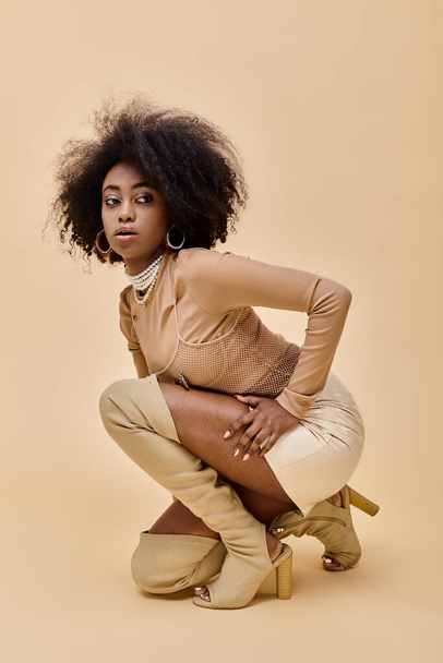 curly african american model in stylish pastel color outfit and thigh-high boots sitting on beige - Фото, зображення