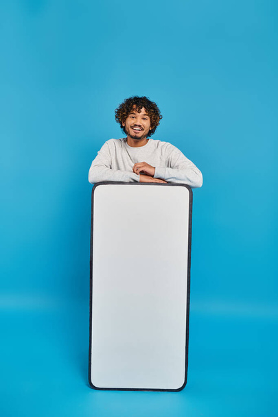 Ένας άντρας στέκεται κρυμμένος πίσω από ένα μεγάλο άσπρο αντικείμενο σε ένα στούντιο πάνω σε ένα μπλε φόντο.. - Φωτογραφία, εικόνα
