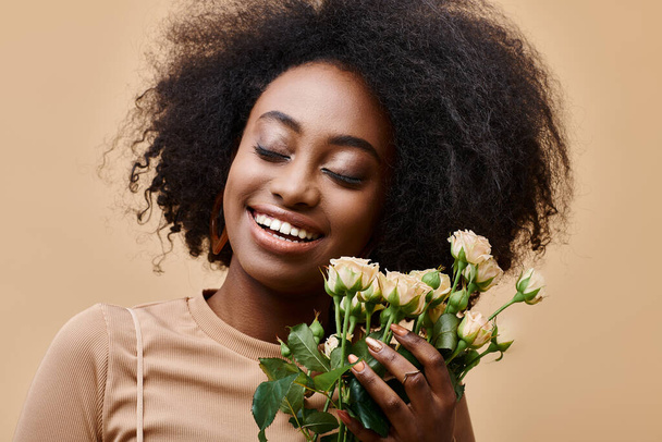 joyful curly african american woman holding tiny roses on beige background, peach fuzz color - Photo, Image