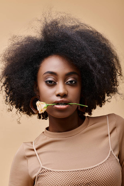 young curly african american woman with small rose in teeth on beige background, peach fuzz - Photo, Image