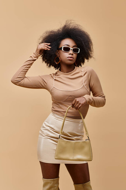 νέο και κομψό αφρικανικό μοντέλο σε γυαλιά ηλίου κρατώντας μοντέρνα τσάντα σε μπεζ φόντο - Φωτογραφία, εικόνα