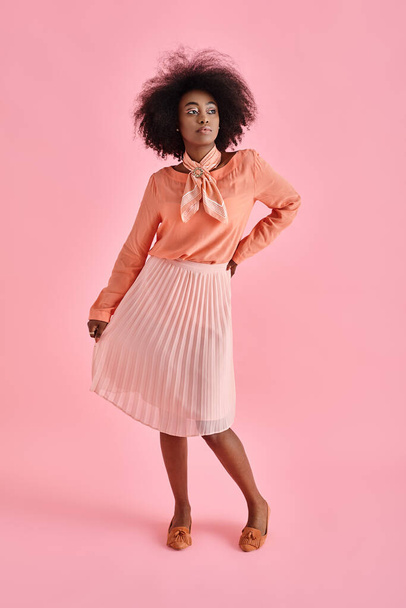 elegante mujer afroamericana en blusa de melocotón y falda midi posando sobre fondo rosa pastel - Foto, Imagen