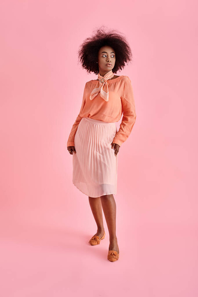 elegancka Afroamerykanka w brzoskwiniowej bluzce i spódnicy midi pozowanie na pastelowym różowym tle - Zdjęcie, obraz