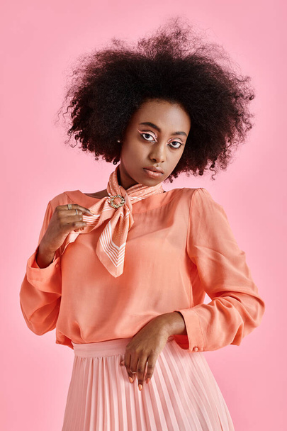 νεαρή Αφροαμερικανή με ροδακινί μπλούζα που προσαρμόζει το κασκόλ του λαιμού και ποζάρει σε ροζ φόντο - Φωτογραφία, εικόνα