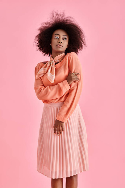 curly african american woman in peach blouse, midi skirt and neck scarf posing on pink background - Photo, Image