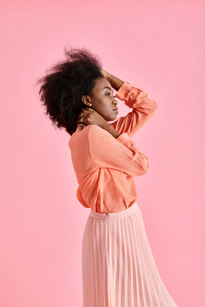 side view, african american woman in peach blouse, midi skirt and neck scarf posing on pink backdrop - Photo, Image