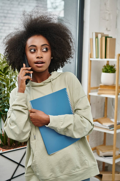 czarna kobieta o zróżnicowanym tle rozmawia przez telefon komórkowy trzymając folder w nowoczesnej przestrzeni dla współpracowników. - Zdjęcie, obraz