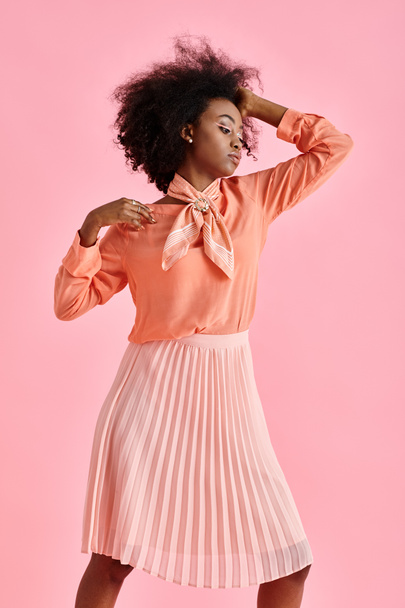 african american woman in peach blouse, midi skirt and scarf adjusting curly hair on pink backdrop - Foto, immagini
