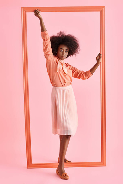 young african american woman in peach blouse and midi skirt posing near frame on pink backdrop - Foto, imagen