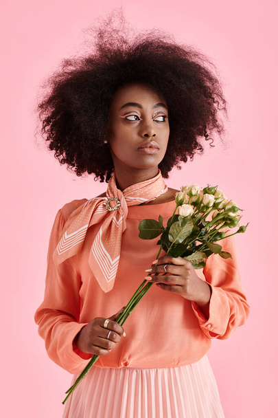 chica afroamericana soñadora en blusa de pelusa de melocotón sosteniendo flores y mirando hacia otro lado en el telón de fondo rosa - Foto, Imagen