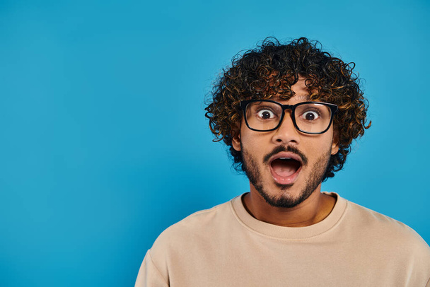 Ένας Ινδός φοιτητής με σγουρά μαλλιά και γυαλιά φαίνεται έκπληκτος σε ένα μπλε φόντο. - Φωτογραφία, εικόνα