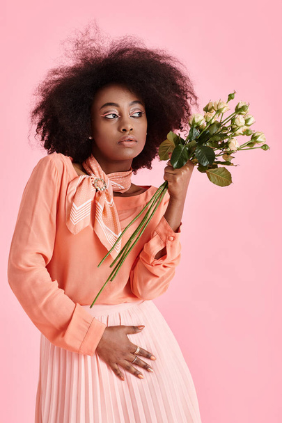 Şeftali tüyü giyinmiş, elinde çiçeklerle pembe arka plana bakan Afro-Amerikan kadın. - Fotoğraf, Görsel