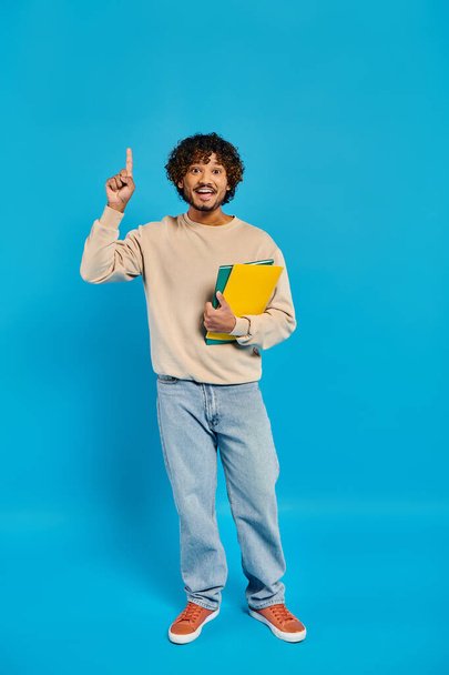 An Indian student in casual attire stands against a blue backdrop, holding a book in one hand and making a peace sign with the other. - Фото, зображення