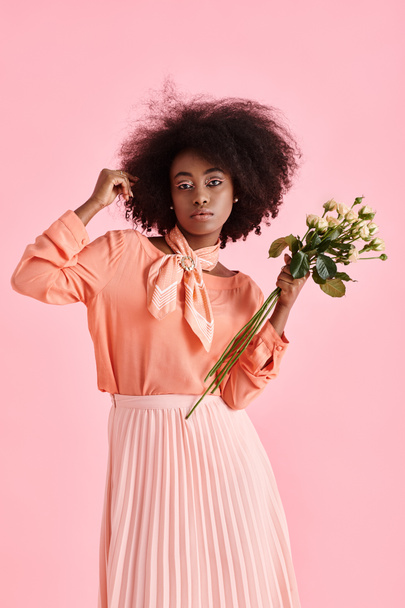 Afrikaans amerikaanse vrouw in perzik fuzz kleding met bloemen en kijken naar camera op roze achtergrond - Foto, afbeelding