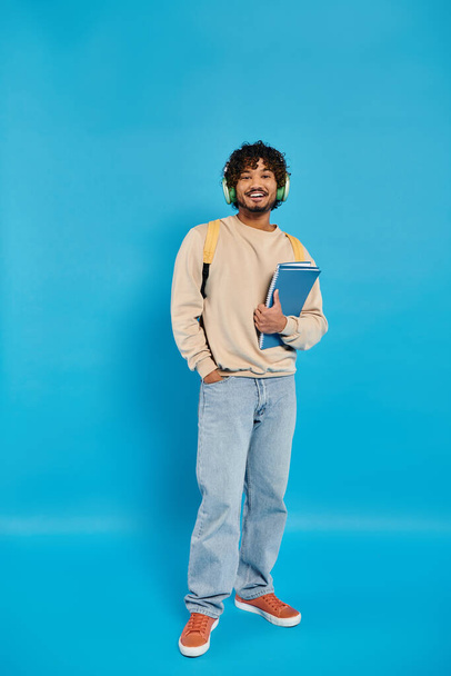 Un estudiante indio de pie con atuendo casual, sosteniendo un libro en sus manos contra un telón de fondo azul en un estudio. - Foto, Imagen