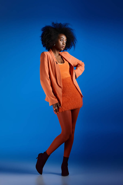 hübsches afrikanisch-amerikanisches Model in strukturellem Kleid und Blazer posiert mit der Hand an der Hüfte auf blau - Foto, Bild