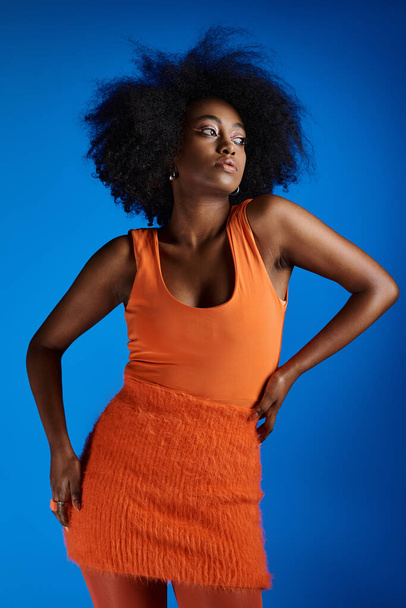 curly african american woman in textured dress posing with hand on hip on vivid blue backdrop - 写真・画像