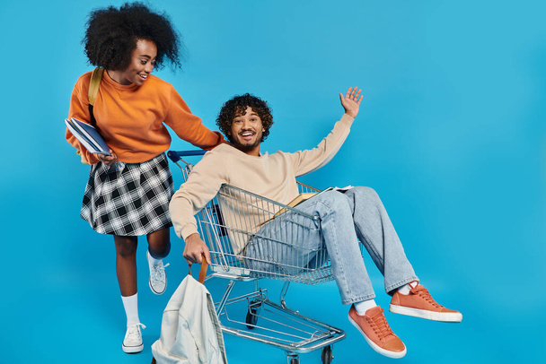 Ein verspieltes Paar gemischtrassiger Studenten in lässiger Kleidung, die zusammen in einem Einkaufswagen sitzen und einen lustigen Moment genießen. - Foto, Bild
