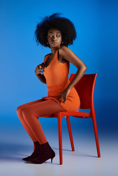 půvabná africká americká žena v oranžových šatech a vysoké podpatky sedí na židli na modrém pozadí - Fotografie, Obrázek