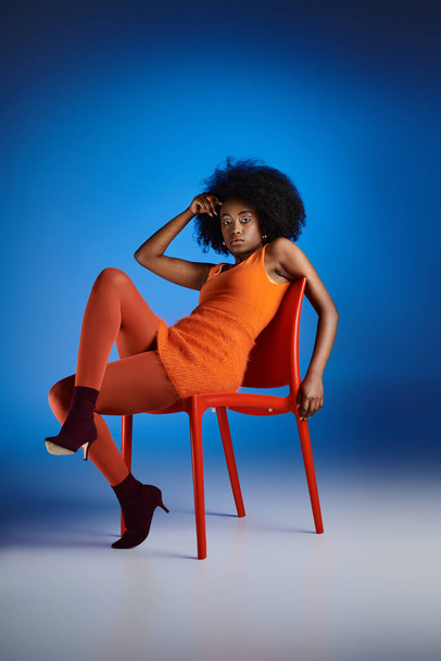 стильная африканская американка в оранжевом платье и на высоких каблуках сидит на стуле на синем фоне - Фото, изображение