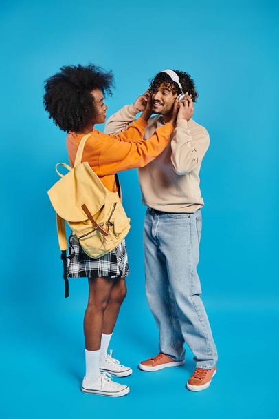 Un hombre y una mujer, ambos estudiantes interracial, de pie juntos en atuendo casual, con la mujer usando una mochila, en un contexto azul. - Foto, Imagen