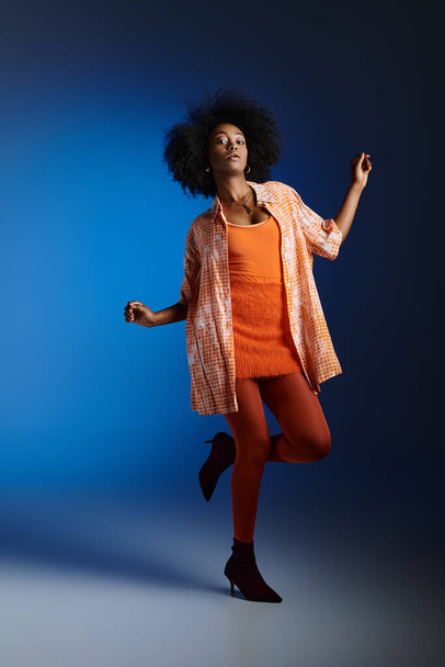 Chic look of african american model in patterned shirt and orange dress posing on blue background - Photo, Image