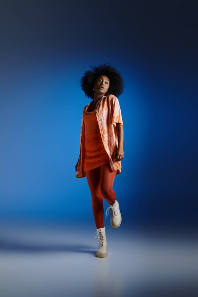 trendy look of african american model in patterned shirt and orange dress posing on blue backdrop - Photo, Image