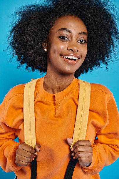 Una alegre universitaria afroamericana con un peinado afro sonríe mientras sostiene un par de tirantes, de pie con atuendo casual contra un telón de fondo azul en un estudio. - Foto, imagen