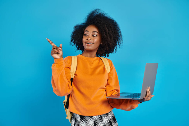 Ein afroamerikanisches College-Mädchen in lässiger Kleidung hält einen Laptop in der Hand und zeigt zur Seite in einem Studio mit blauem Hintergrund. - Foto, Bild