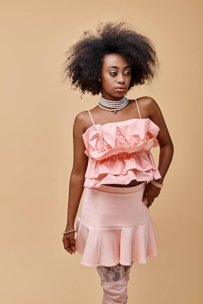 curly young african american woman in peach ruffled top and mini skirt posing on beige background - Photo, Image
