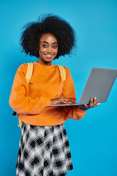 Afroamerykańska studentka uśmiecha się trzymając laptopa w studio na niebieskim tle. - Zdjęcie, obraz