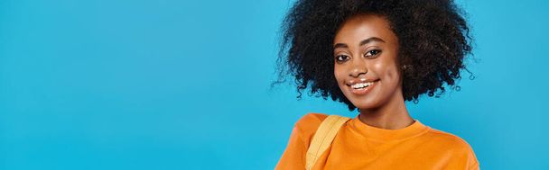 Uma menina da faculdade com um grande afro sorri calorosamente para a câmera contra um pano de fundo azul em um estúdio. - Foto, Imagem