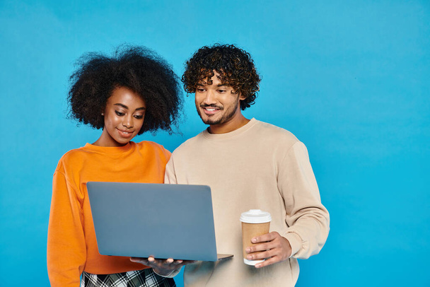 Un homme et une femme, tenant un ordinateur portable et une tasse de café, debout ensemble dans un studio avec une toile de fond bleue. - Photo, image
